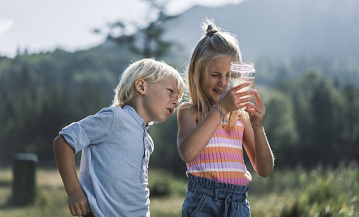 Zwei Kinder in Grünberg im Salzkammergut begutachten ein mit Wasser gefülltes Einmachglas. Im Hintergrund ist undeutlich die gründe Landschaft zu sehen.